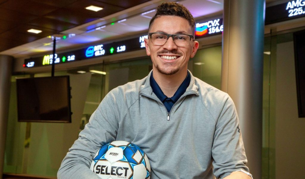 Photo of Nick Kinzler holding a soccer ball in front of the stock ticker in the Simms Center